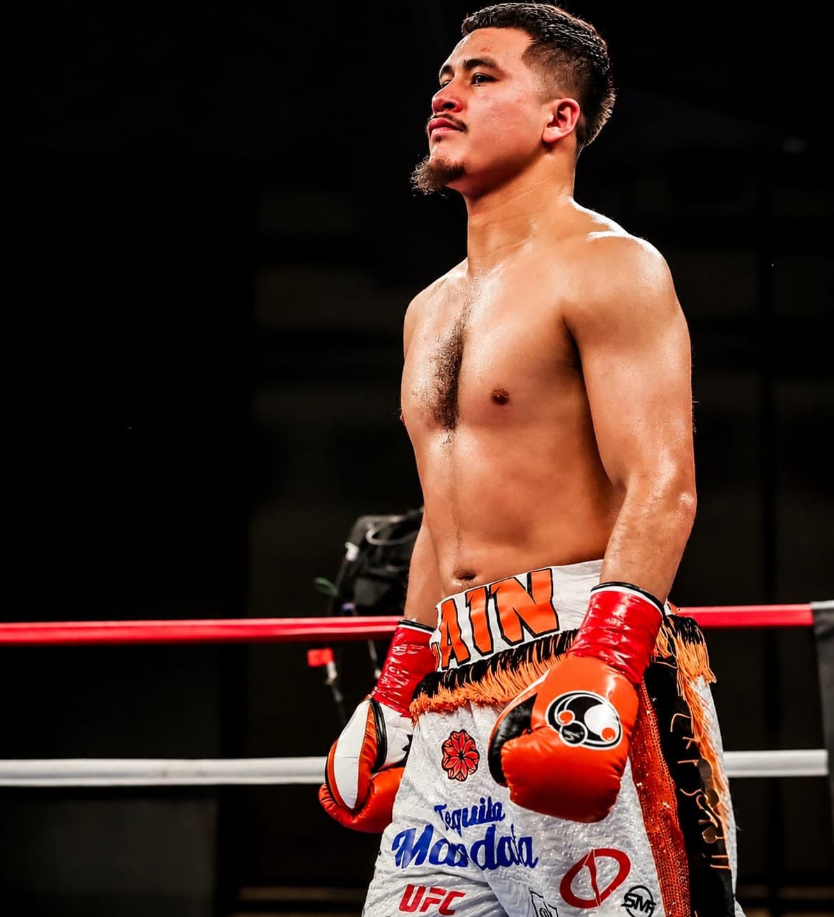 A boxer standing in the ring after a bout, hands in orange gloves, looking up under the lights.
