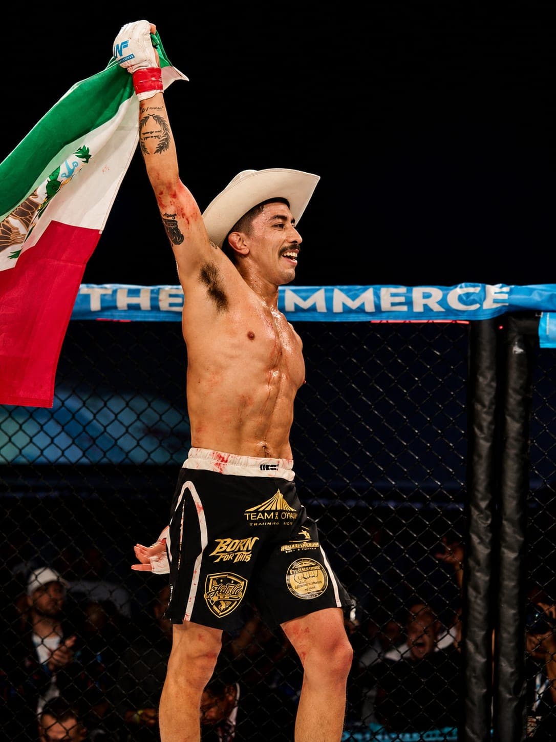 A fighter in a white hat raising a Mexican flag over his head inside the cage, fans behind the fence.